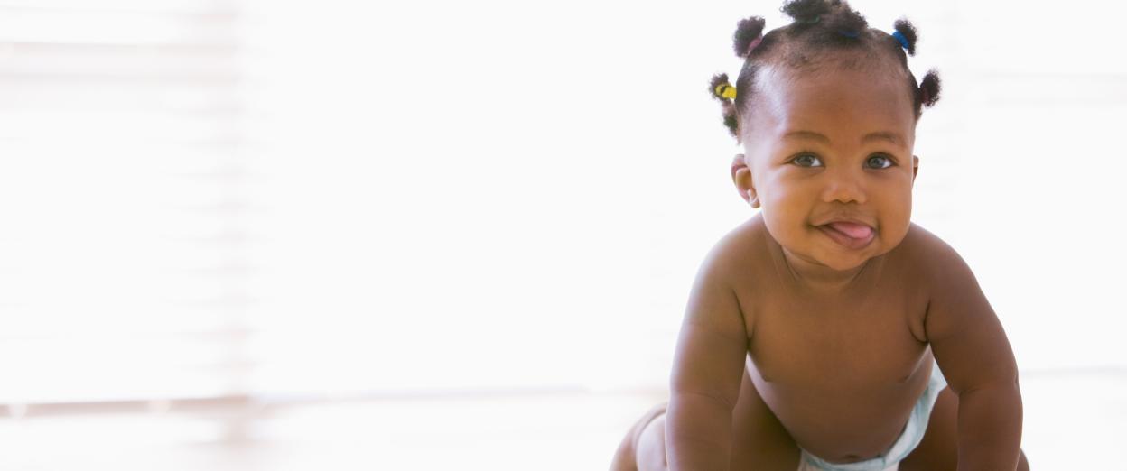10 month old baby girl with dark skin tone and tiny pigtails crawling towards camera with tongue out