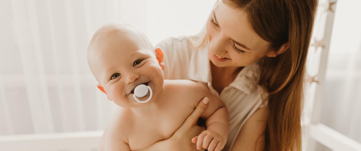 11 Month Old baby with almost no hair smiling with a pacifier in their mouth while being held by mom. Baby is wearing a diaper and mom is smiling at the baby lovingly