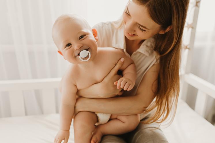11 Month Old baby with almost no hair smiling with a pacifier in their mouth while being held by mom. Baby is wearing a diaper and mom is smiling at the baby lovingly