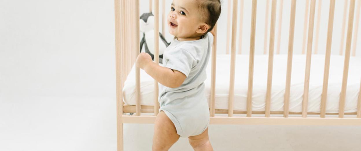 12 month old baby learning to stand holding on to his crib. Baby has olive complexion and dark hair and eyes. He is looking proudly at someone off camera