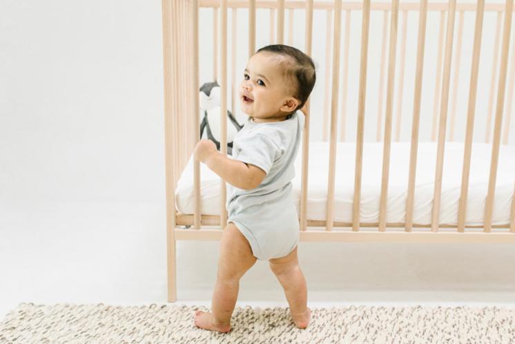 12 month old baby learning to stand holding on to his crib. Baby has olive complexion and dark hair and eyes. He is looking proudly at someone off camera