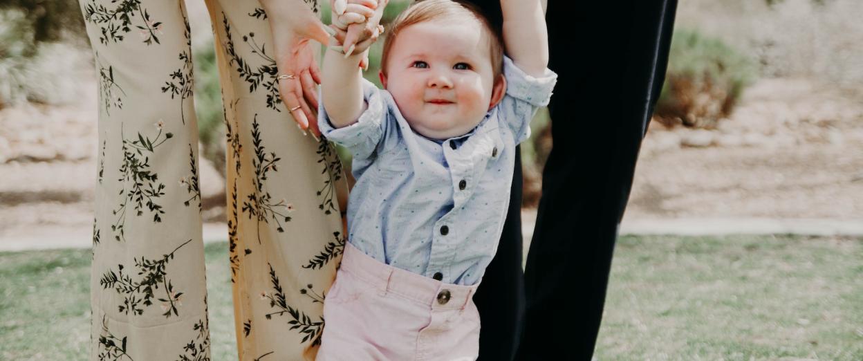 14 month old boy holding 2 adult's hands (the adults are cut off at the waist) while learning to walk. The baby has a light skin tone and reddish hair.
