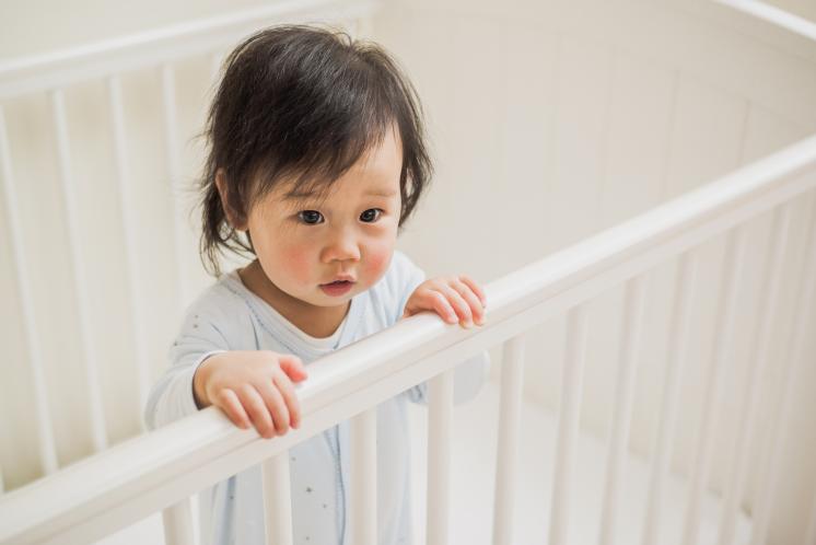 14month old toddler standing in crib. Baby is Asian. They look like they just woke up or have been crying with puffy under eyes and slightly messy dark hair