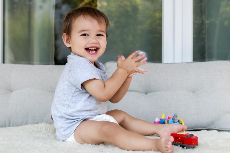 16 month old toddler sitting on a fuzzy surface while playing with toys and clapping hands. The baby has a medium skin tone and brown hair. He has a red train at his feet and is wearing a grey shirt and a diaper