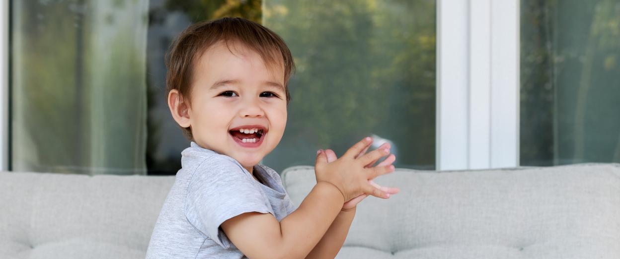 16 month old toddler sitting on a fuzzy surface while playing with toys and clapping hands. The baby has a medium skin tone and brown hair. He has a red train at his feet and is wearing a grey shirt and a diaper