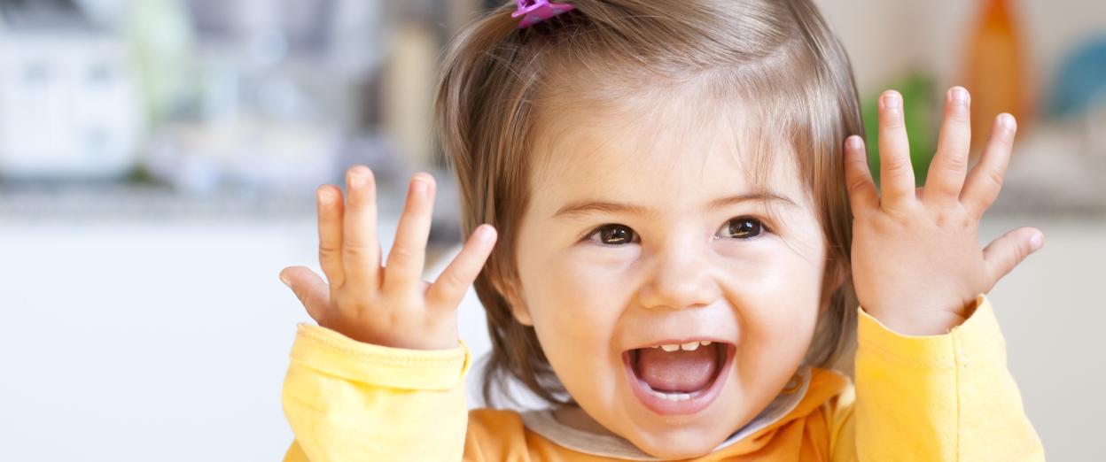 20 month old girl smiling at the camera with her hands up by her face