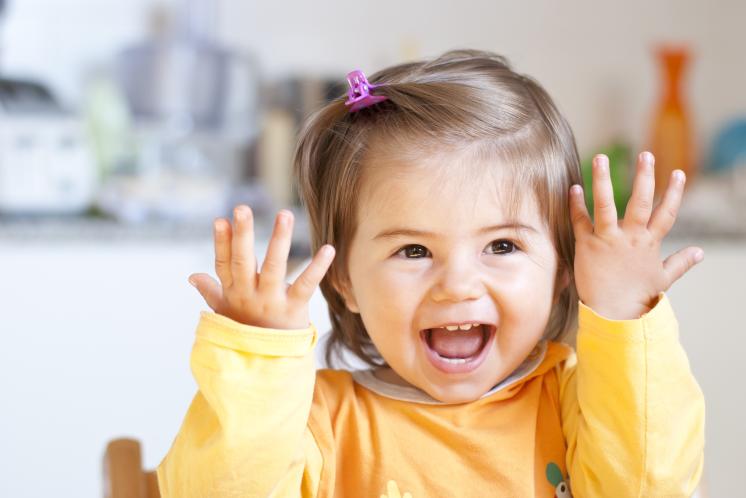 20 month old girl smiling at the camera with her hands up by her face