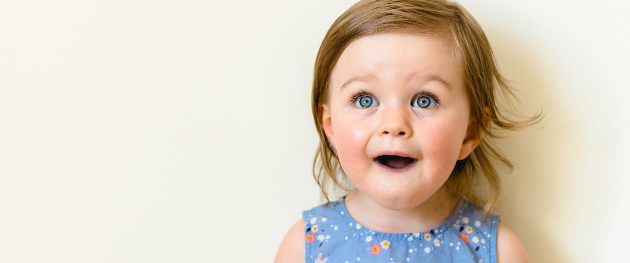 Older baby girl wearing blue tank top with light colored flowers looking up at the camera with a surprised face