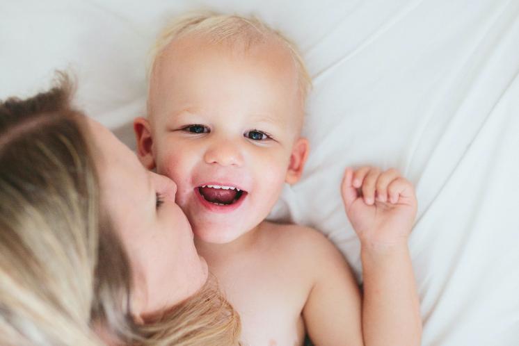 happy 2 year old on bed with blonde mom kissing his cheek