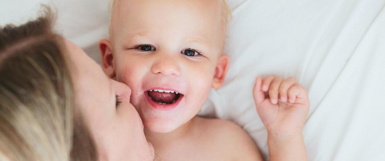 happy 2 year old on bed with blonde mom kissing his cheek