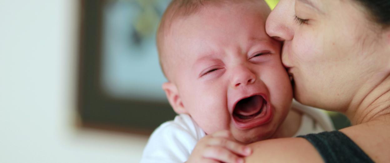 Baby fighting sleep and crying on mom's shoulder while she gives him a kiss