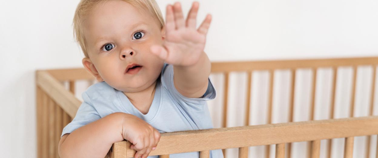 baby standing in crib reaching toward camera while refusing to nap