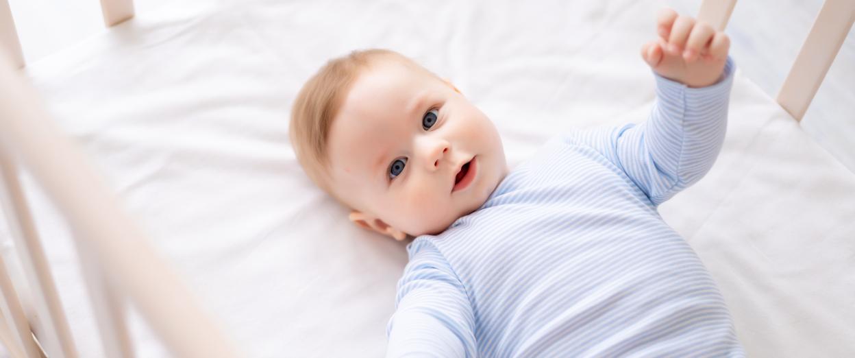 Baby boy wearing a blue striped onesie looking at that camera from inside his crib
