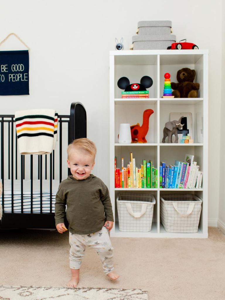 toddler boy standing in the middle of the nursery with book shelves full of colorful books and toys