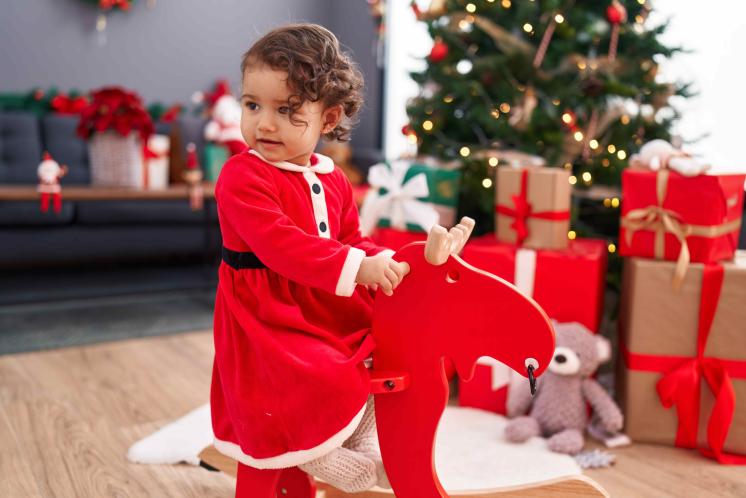 toddler girl sitting on red rocking horse in front of christmas tree wearing red santa themed dress