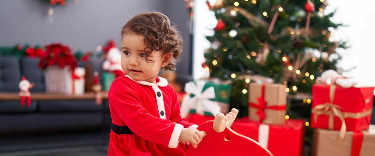 toddler girl sitting on red rocking horse in front of christmas tree wearing red santa themed dress