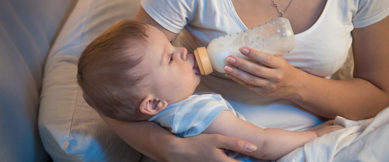 woman sitting on bed cross legged with baby across her lap while she feeds her baby with a bottle of forumla