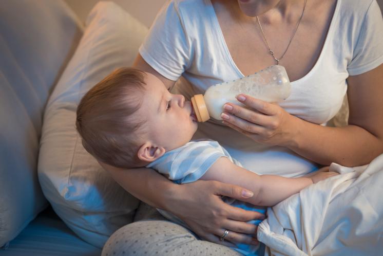 woman sitting on bed cross legged with baby across her lap while she feeds her baby with a bottle of forumla