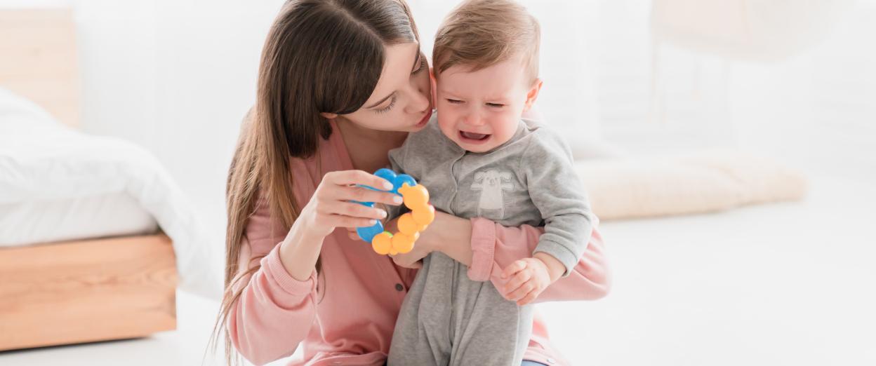 Mom kneeling down trying to calm overtired baby by shushing and holding out a blue and yellow rattle.