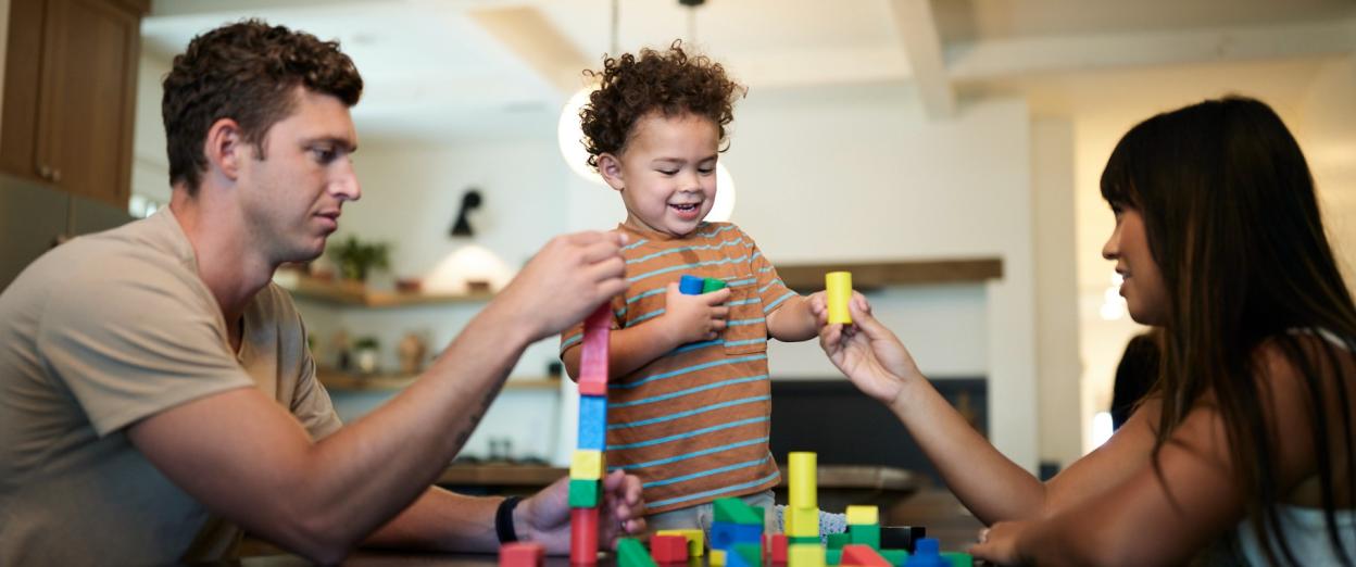 man, 2 year old toddler smiling, and woman playing with blocks at a table