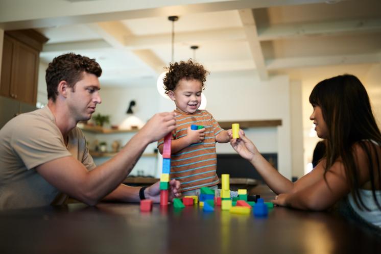 man, 2 year old toddler smiling, and woman playing with blocks at a table