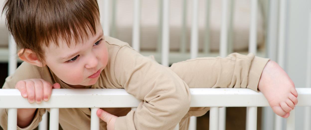 toddler in neutral pajamas lifting leg over the side of a white crib to climb out
