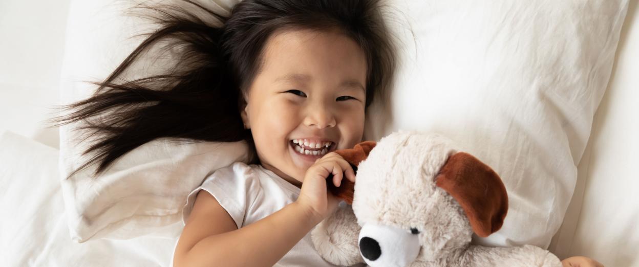 Toddler girl laying in white sheeted bed smiling at camera while holding a stuffed animal