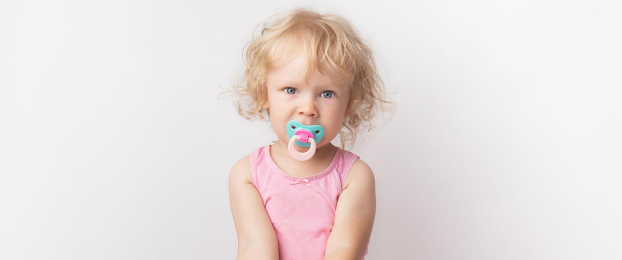 Toddler girl wearing pink tank top looking at the camera with a teal and pink pacifier in her mouth