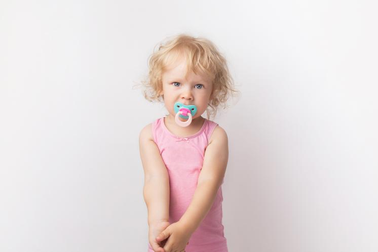 Toddler girl wearing pink tank top looking at the camera with a teal and pink pacifier in her mouth