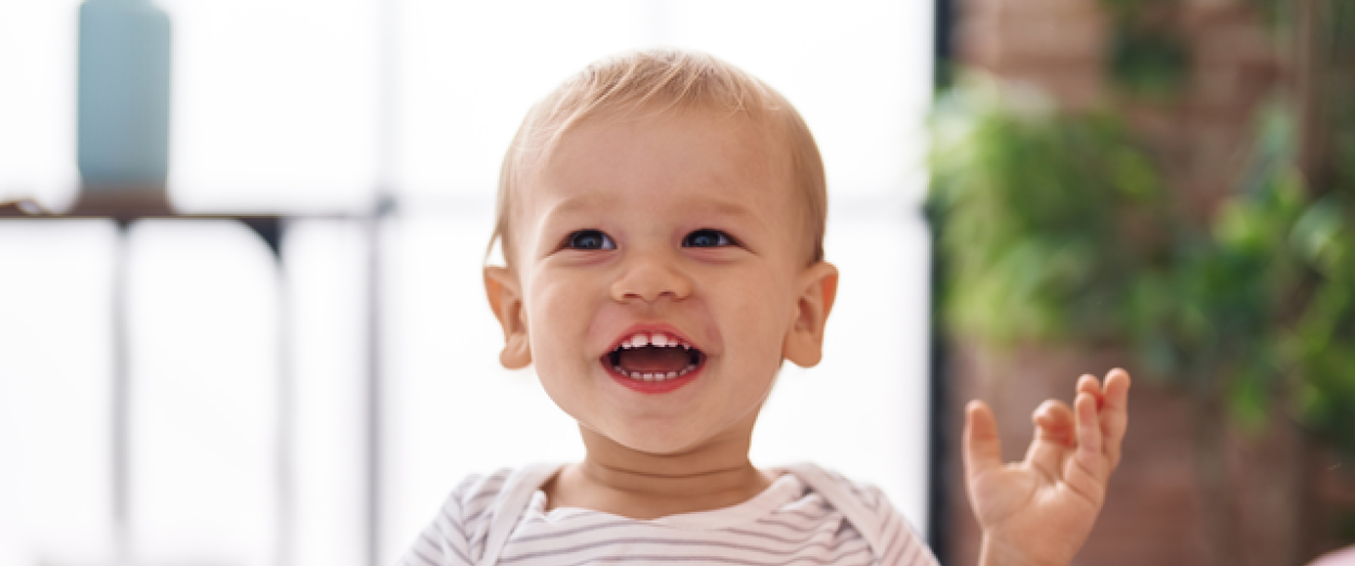 15 month old toddler with a light skin tone blonde hair smiling at the camera with hands up. The child is wearing a striped shirt.