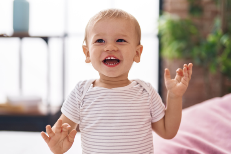 15 month old toddler with a light skin tone blonde hair smiling at the camera with hands up. The child is wearing a striped shirt.
