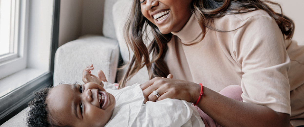 17 month old playing with mom on couch smiling at the camera. Mom and baby have a dark skin tone. Baby has black curly hair and mom has dark brow hair. She is wearing a light beige mock neck shirt and a red bracelet.